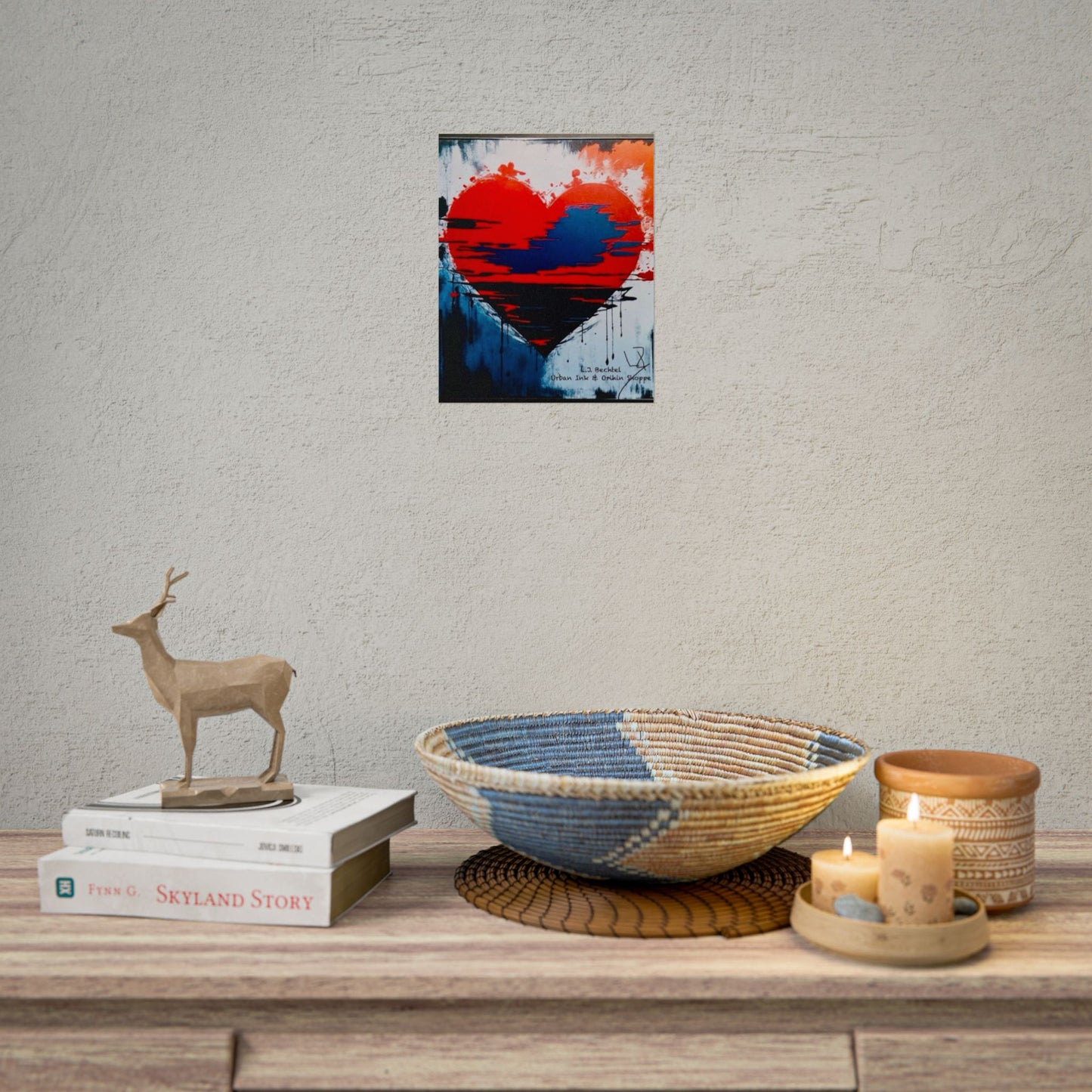 Decorative setup with a heart-shaped painting, books, a deer figurine, a woven bowl, and candles on a wooden surface.