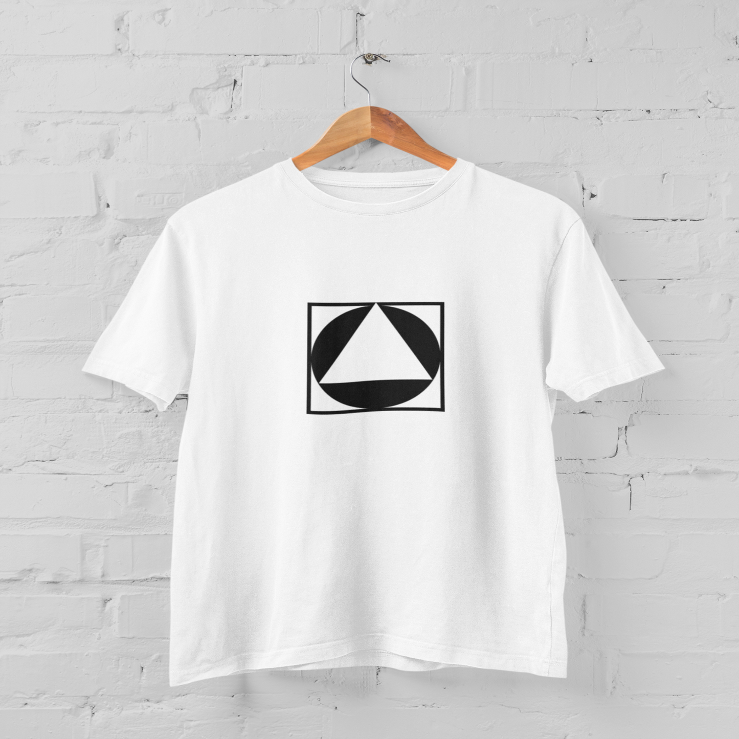 White t-shirt with a black geometric design on a hanger against a white brick wall.